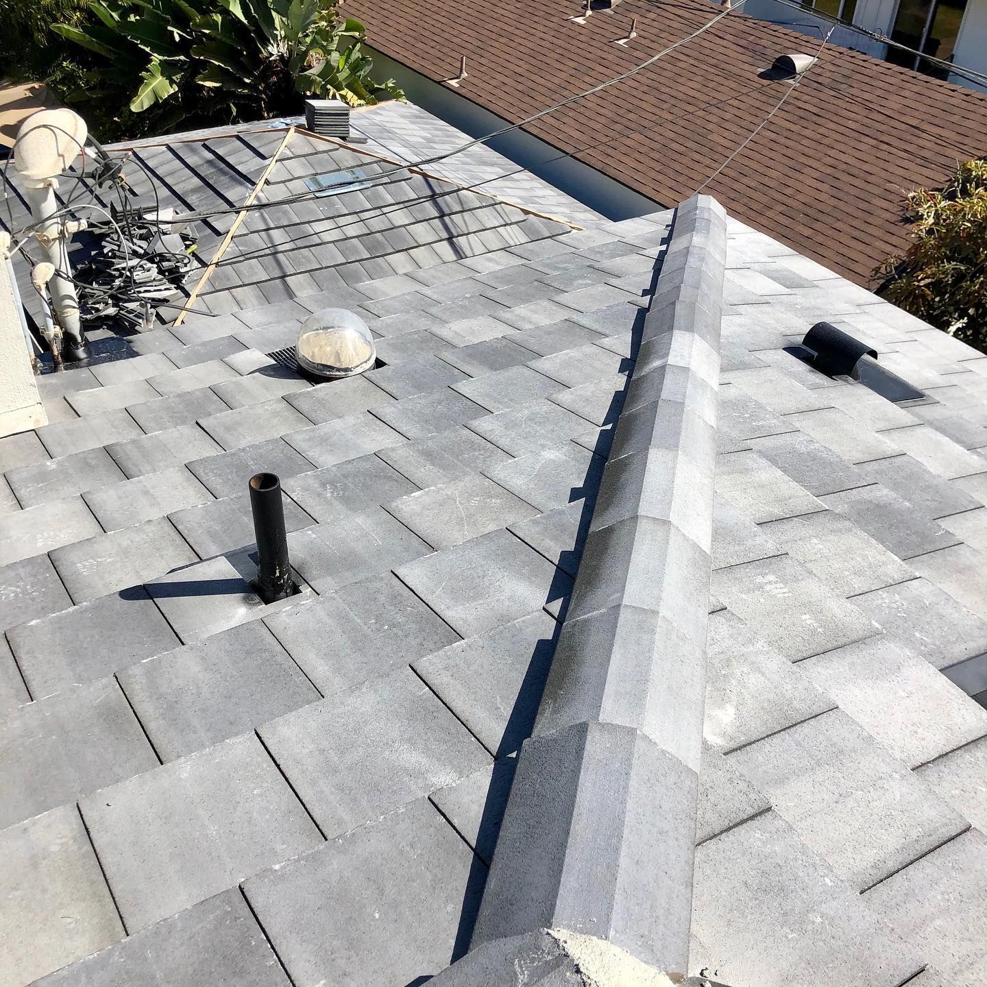 Gray shingle roof with vents and a chimney against a background of other houses and greenery.