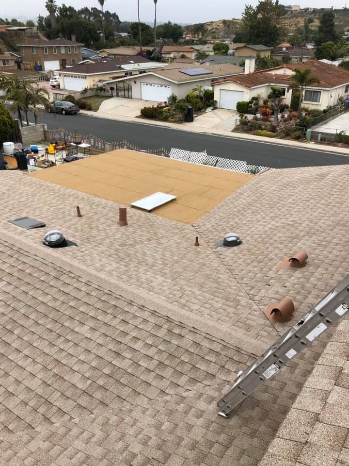 A roof being worked on, partial shingle replacement. Brown shingles, open plywood section. A ladder leans against the roof.