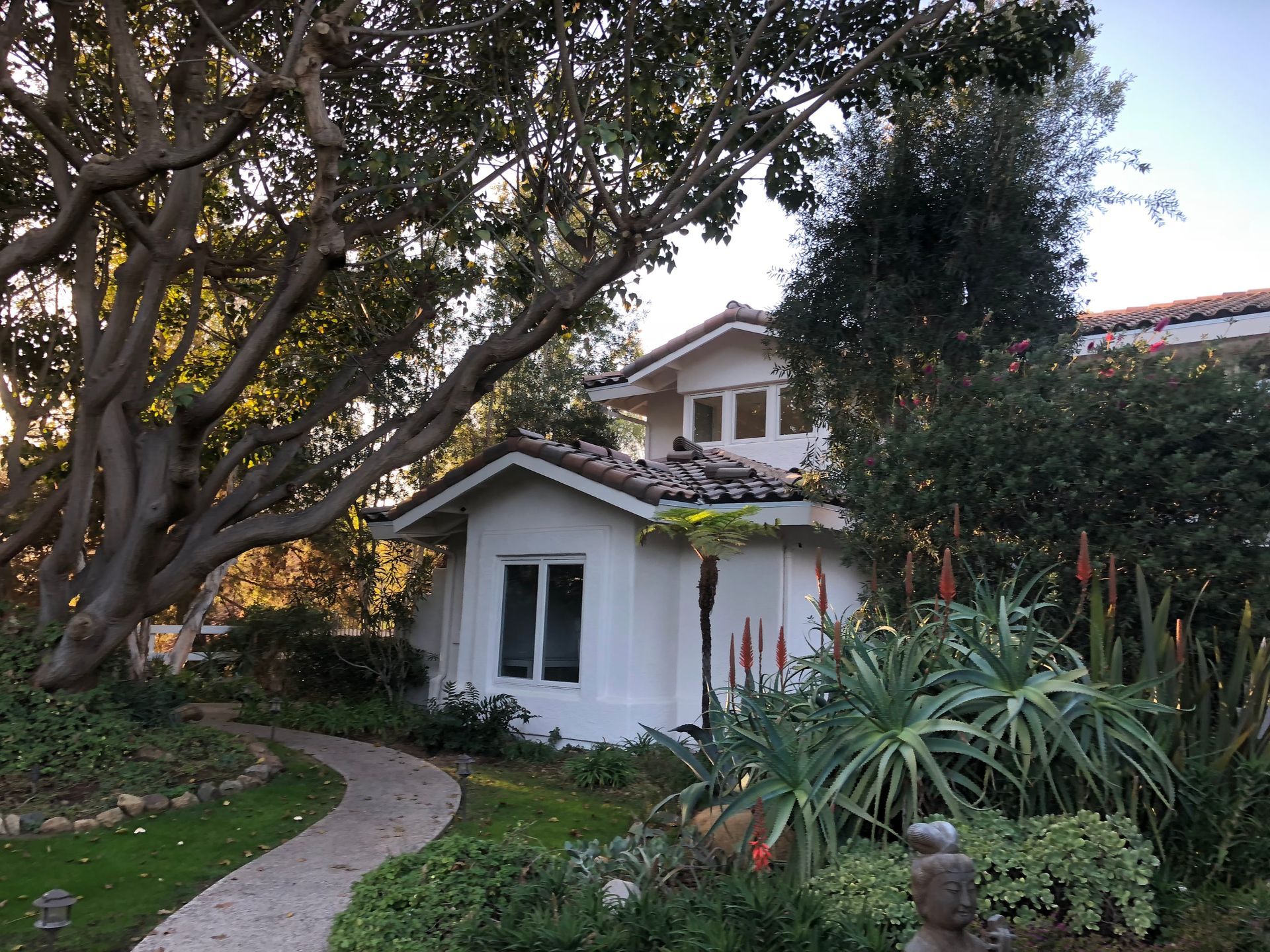 White cottage and garden path leading to a two-story home, shaded by trees and lush greenery.