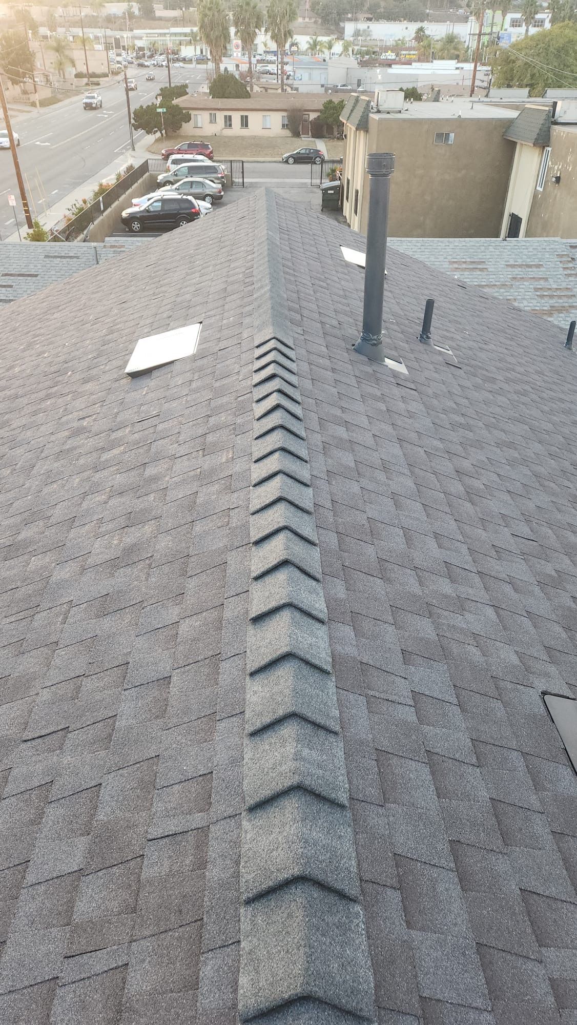 View of a dark asphalt shingle roof with a central ridge cap, chimney, and urban background.