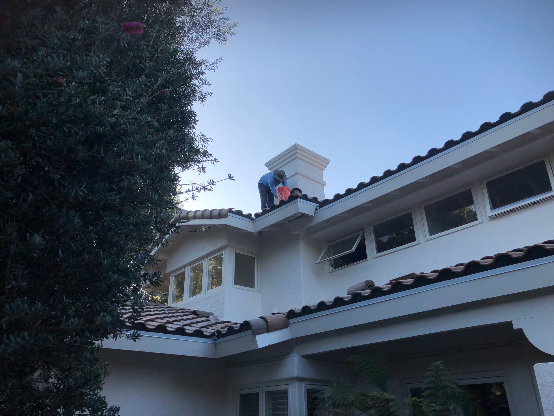 Two people on a roof working near a chimney. White stucco house with tile roof. Blue sky.