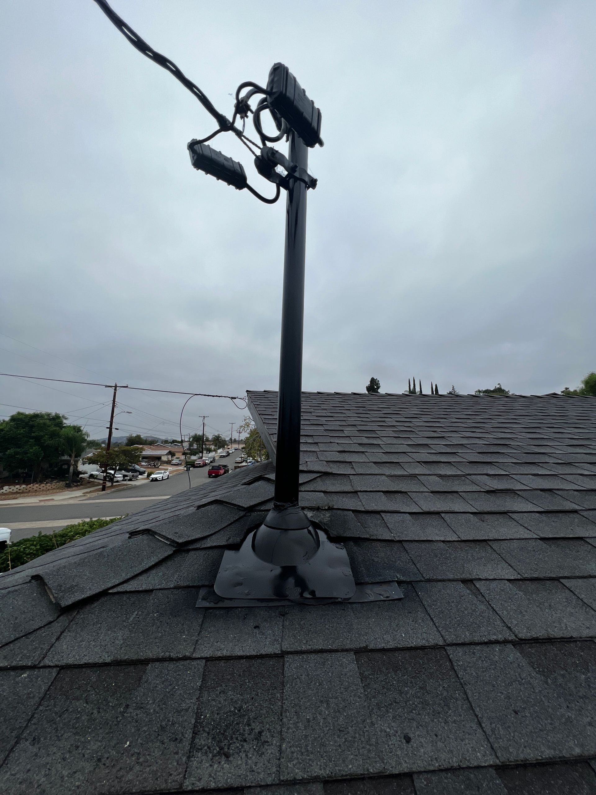 Black utility pole with wires attached, mounted on a dark shingle roof against a cloudy sky.
