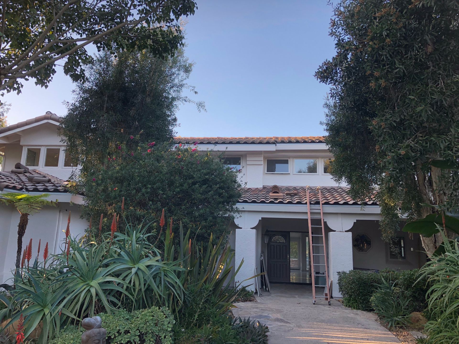 White house with a tiled roof, trees, and landscaping. A ladder leans against the building.