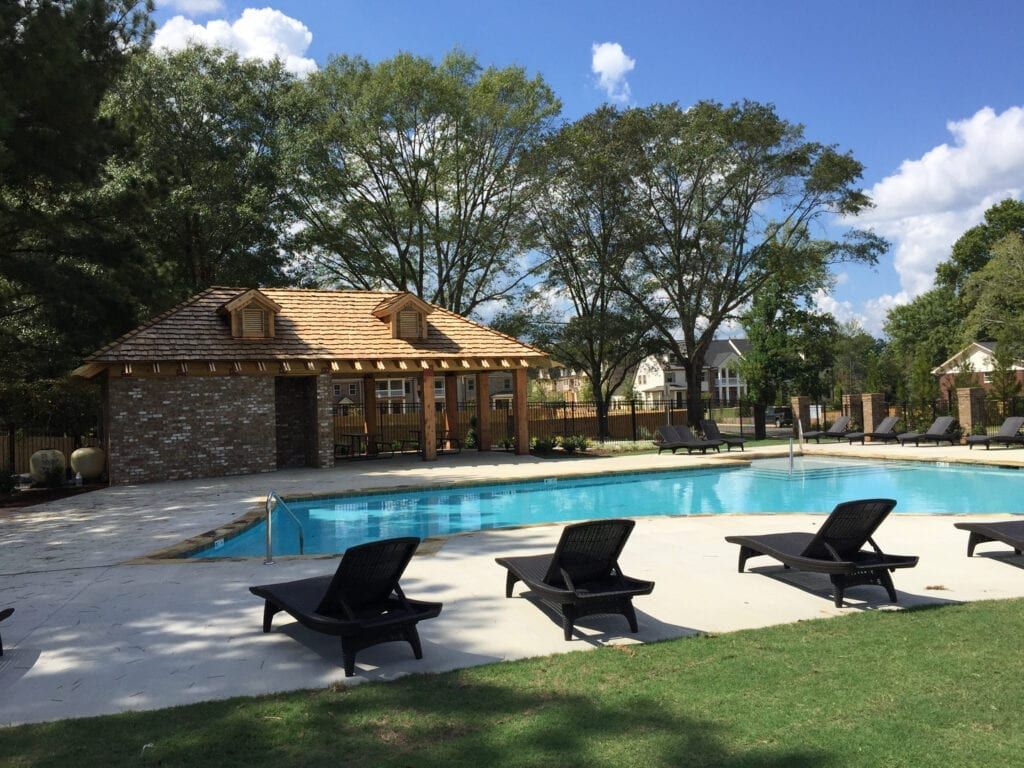 Poolside scene with lounge chairs, a building with a thatched roof, and trees under a blue sky.