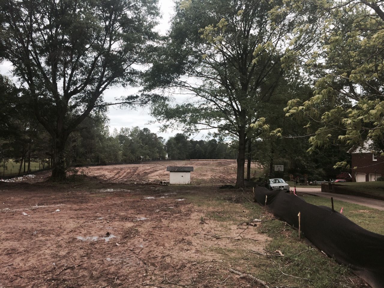 Cleared dirt lot with trees in the background, a white structure, and a black barrier on the right.