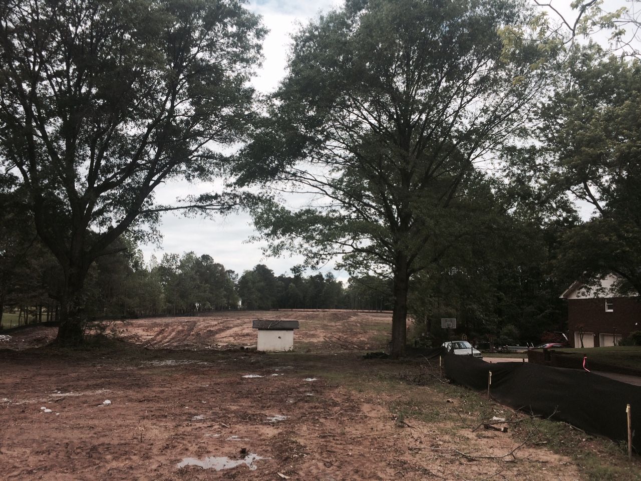 Dirt lot with trees, construction fencing, and utility box under a cloudy sky.