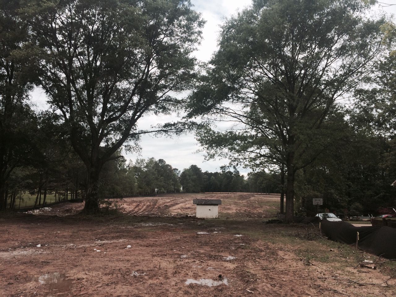 A cleared lot with trees on either side, cloudy sky above. A small concrete structure sits in the center.