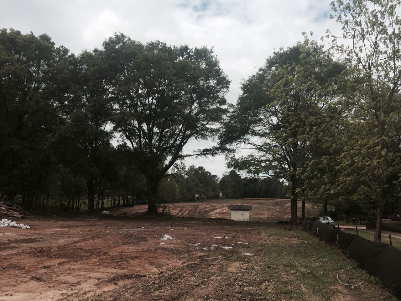 Cleared dirt field with trees and a small structure in the background under a cloudy sky.