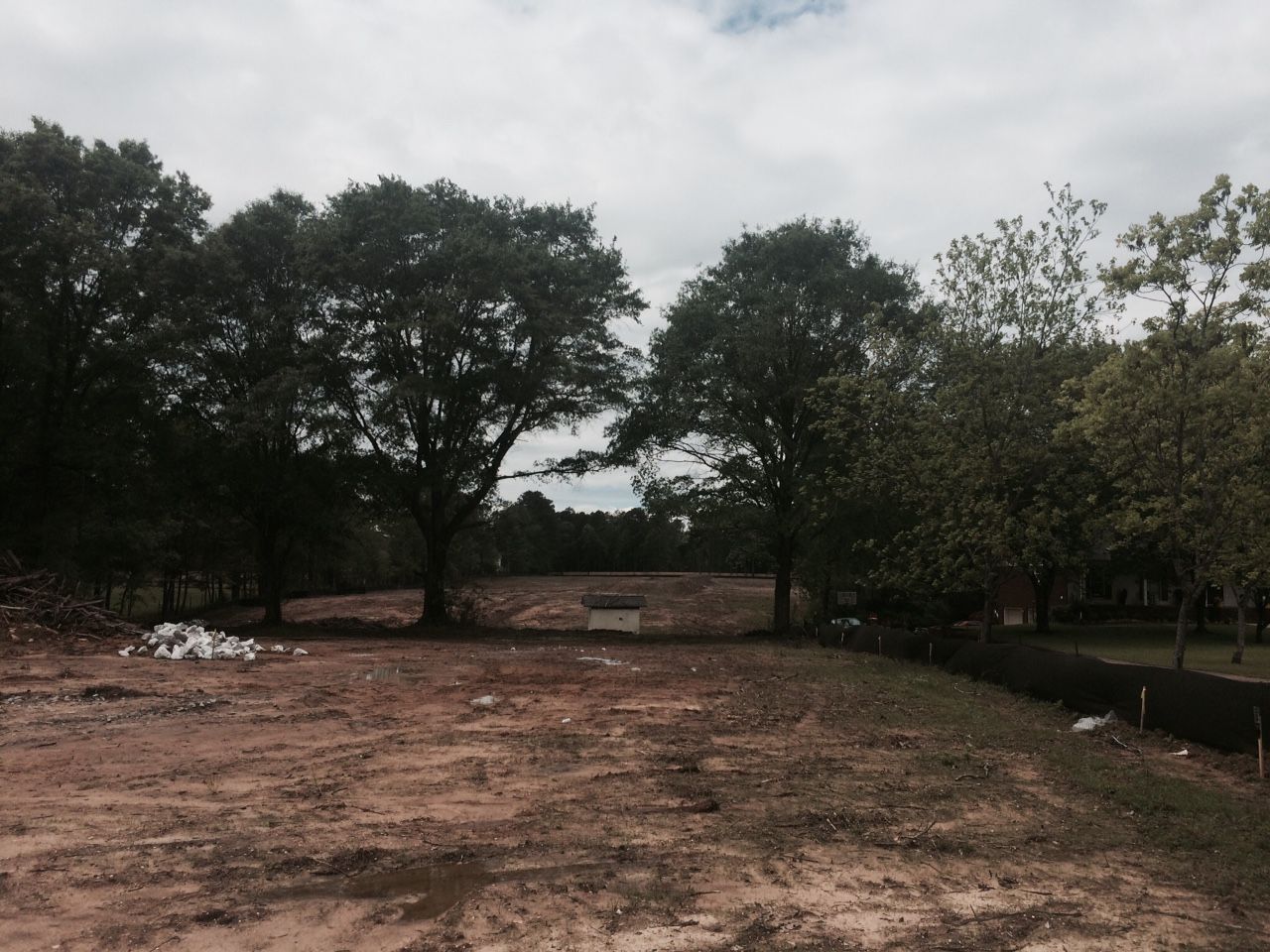 Cleared dirt lot with sparse grass and trees under a cloudy sky.