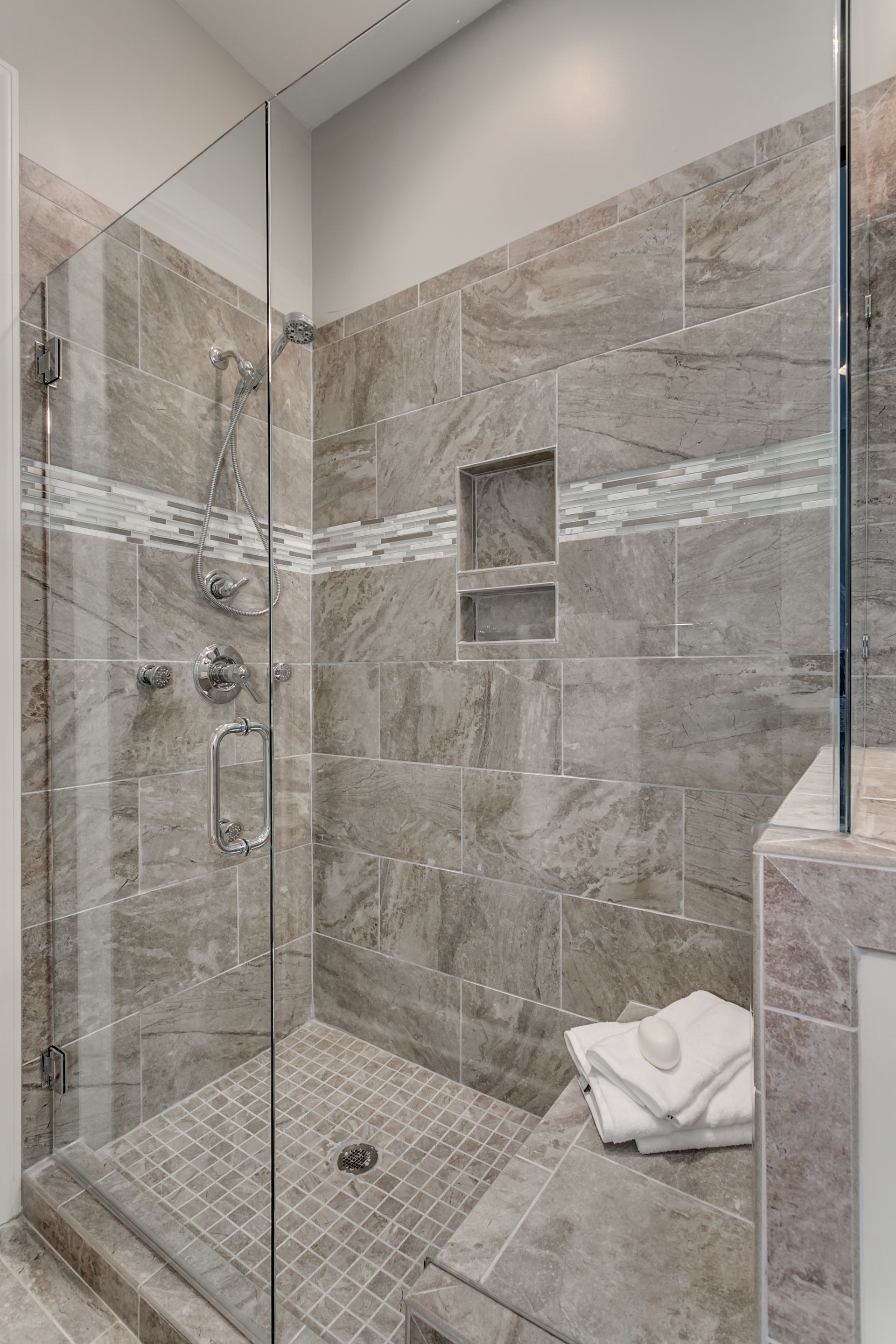 Shower with glass door, gray tiled walls, small mosaic floor, white towels on a bench.