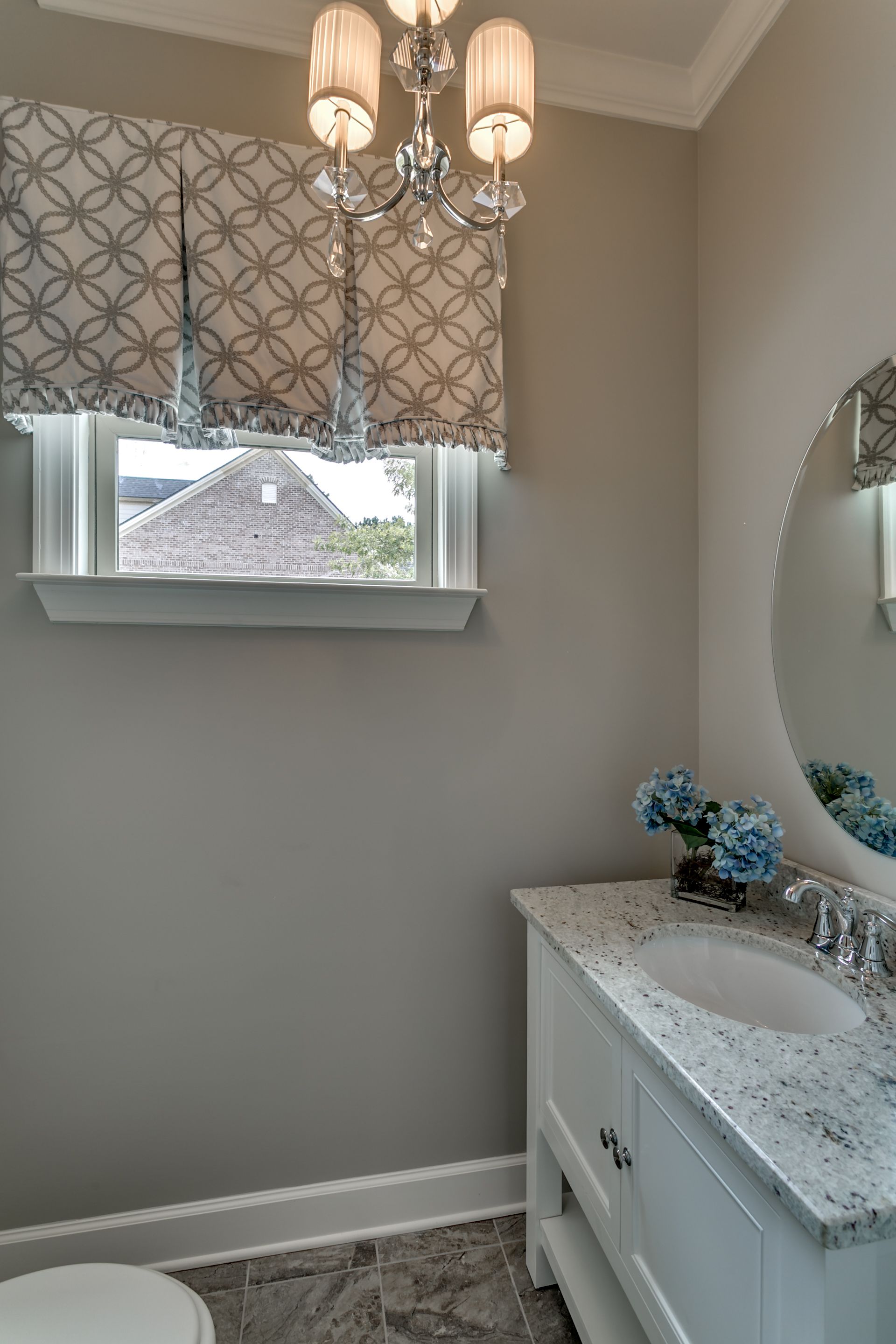 Powder room with grey walls, white vanity, round mirror, and patterned window shade.