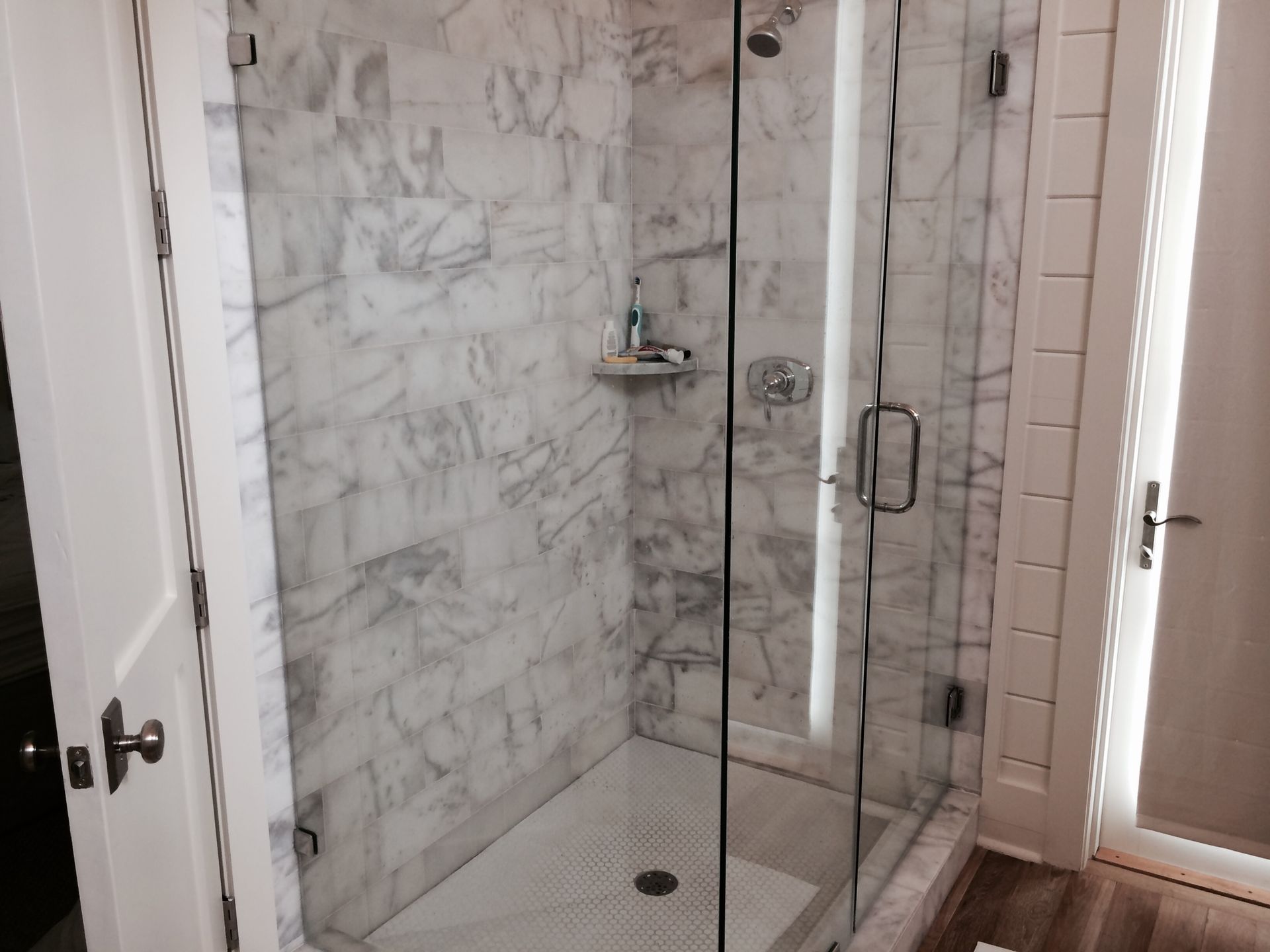 Marble-tiled shower with a glass door. Soap and toiletries on a small shelf.