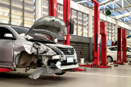 Damaged silver car in an auto repair shop with its hood open and front end crushed.