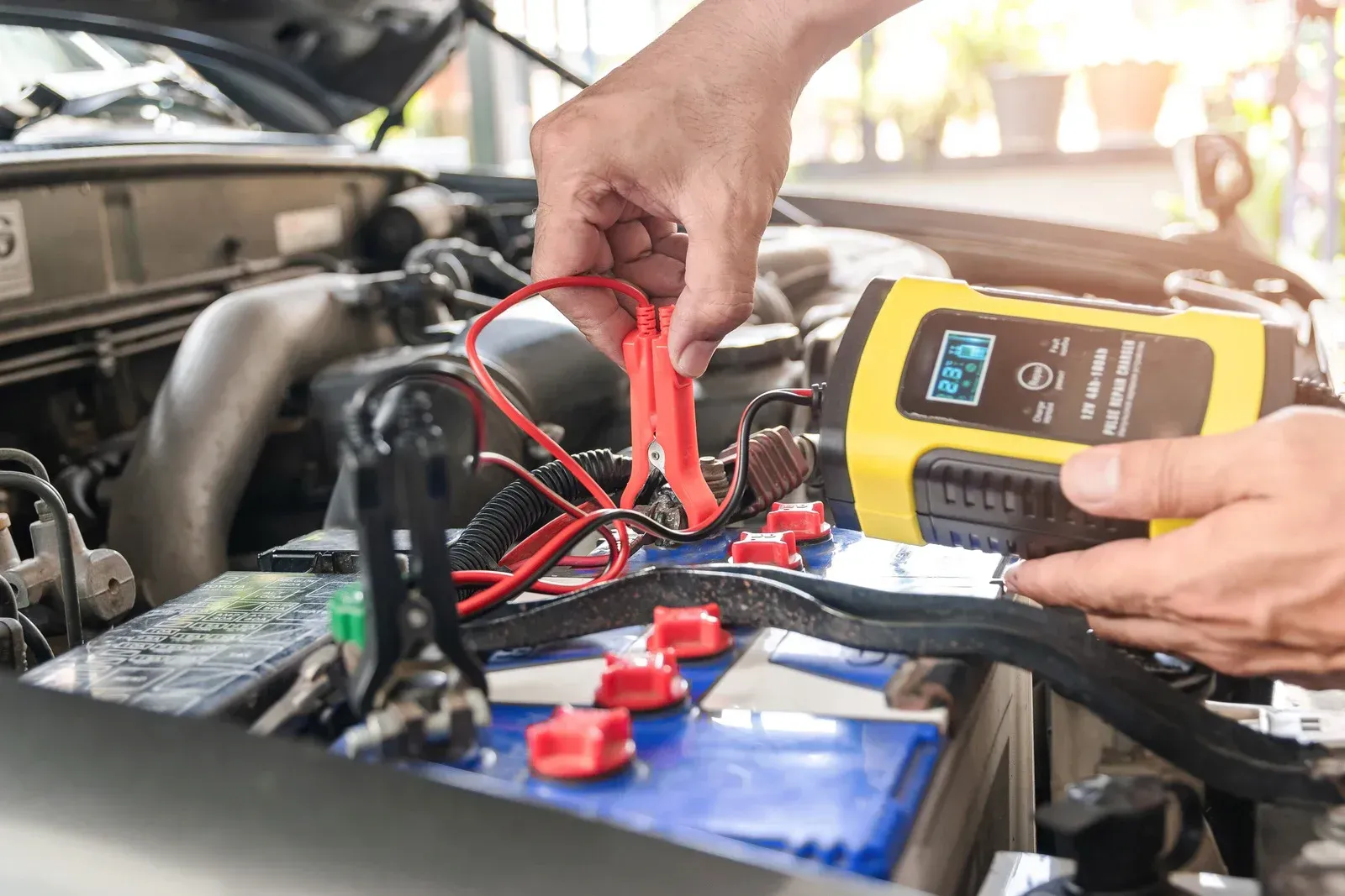 Hands connecting a red clamp to a car battery terminal while using a yellow battery charger in an engine bay.