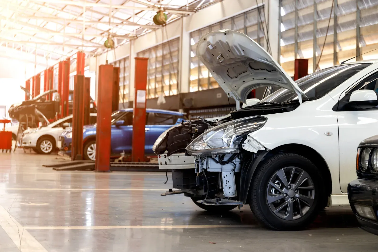 Cars in a repair shop, one with hood open, being serviced.  Light-filled interior.