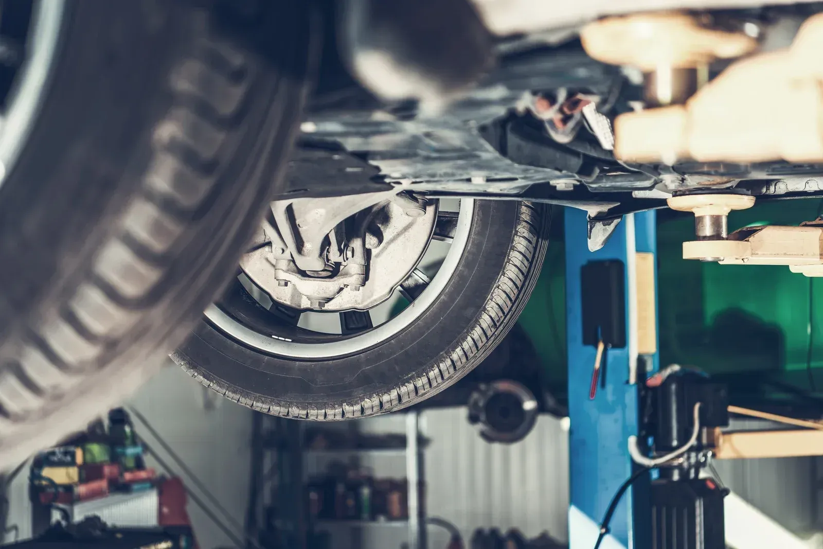 Car on a lift in a repair shop, viewed from underneath. Tire and undercarriage visible.
