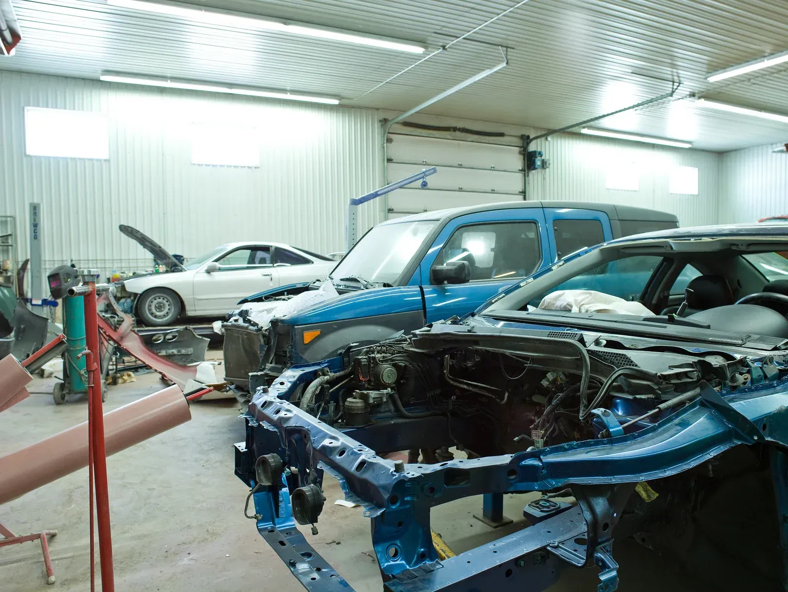 Cars in various states of repair inside a body shop. Blue, white vehicles. Tools and equipment visible.