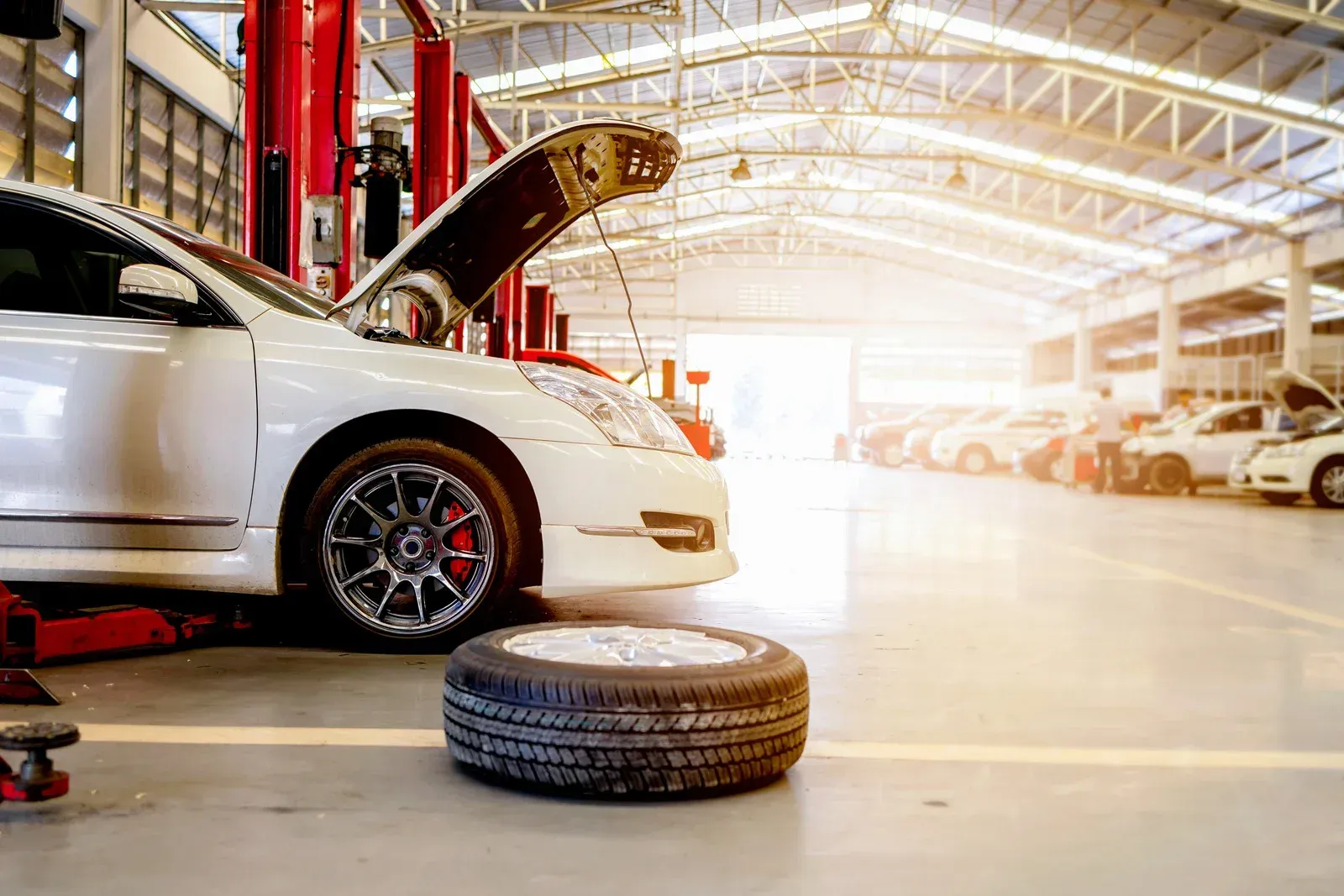 White car in a repair shop, hood open, a tire on the floor, red lift, sun shining.