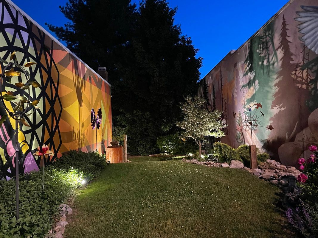 A grassy garden courtyard at twilight, bordered by two walls featuring colorful geometric and forest-themed murals.
