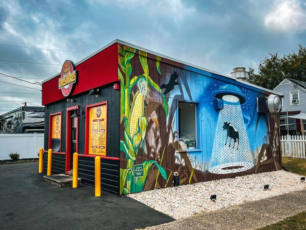 A small, flat-roofed building features a vibrant mural of a UFO abducting a cow in a field under a blue, cloudy sky.