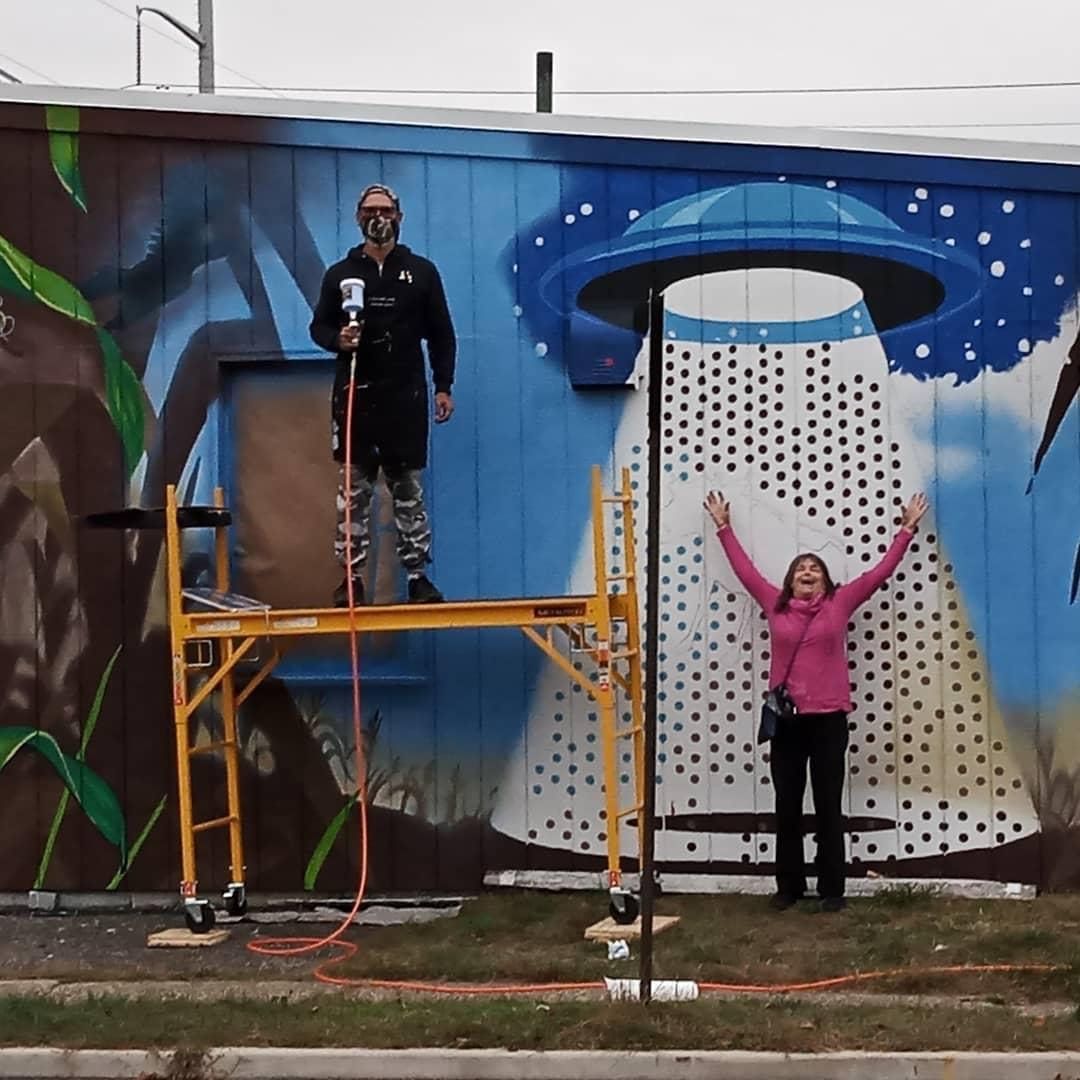 Two people pose in front of a colorful wall mural featuring a large UFO with a light beam, one standing on scaffolding.