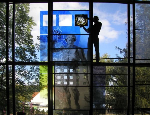 Person cleaning a large window with a squeegee, reflecting a statue and trees outside.
