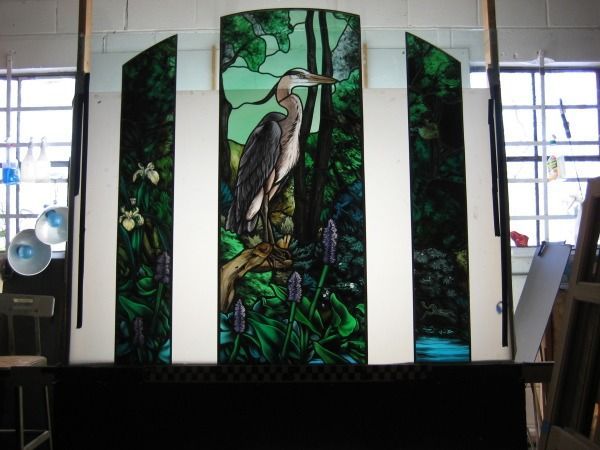 A triptych of stained glass panels featuring a great blue heron perched in a lush, green forest setting.