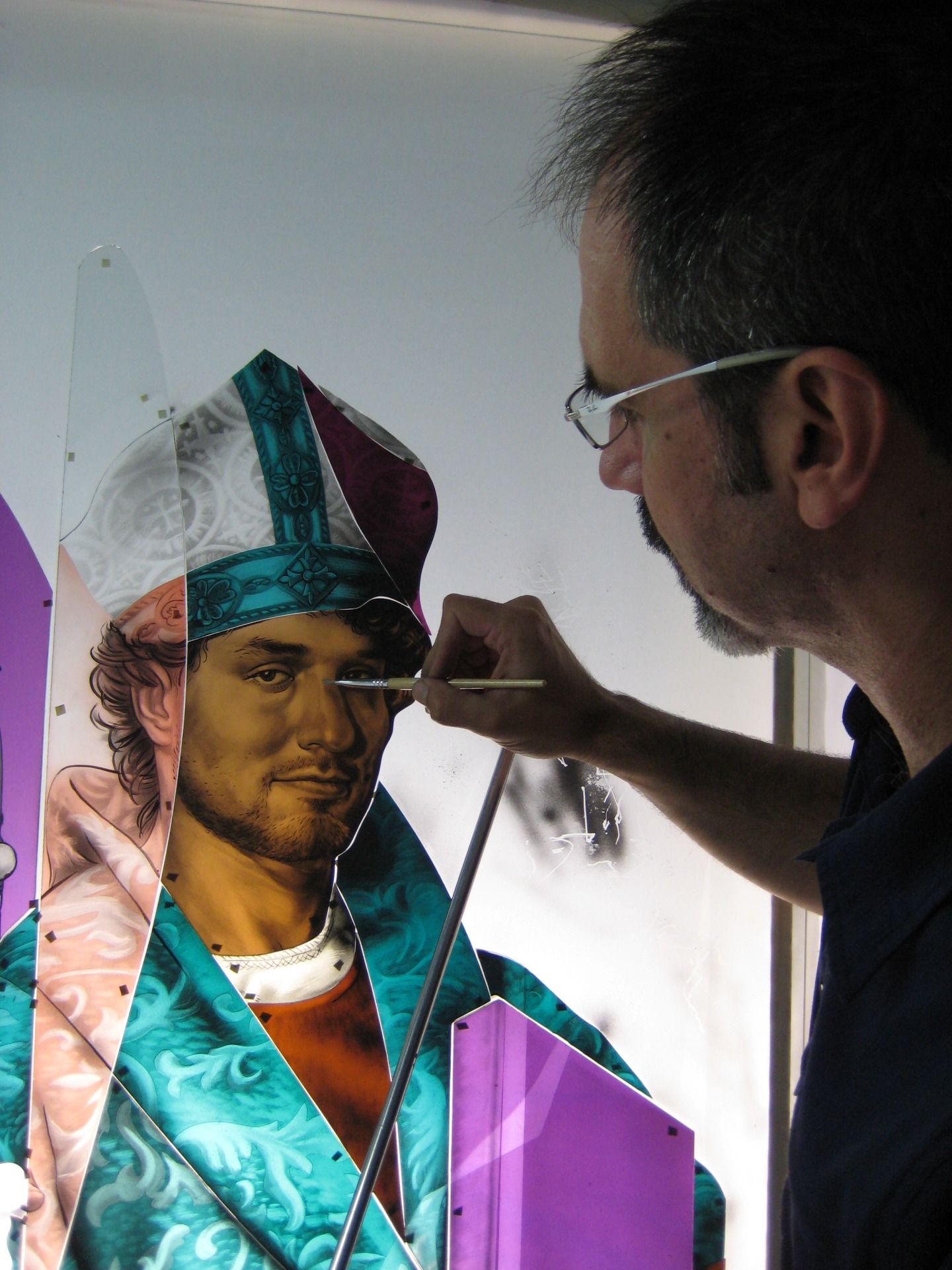 A person meticulously paints the eye of a portrait of a person wearing a formal, decorative hat and teal robes.