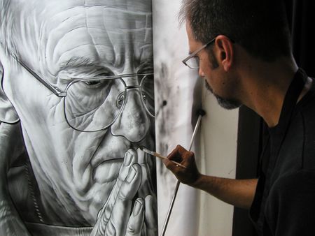 A person in glasses paints a detailed, black-and-white portrait of a man with his hands clasped in prayer.
