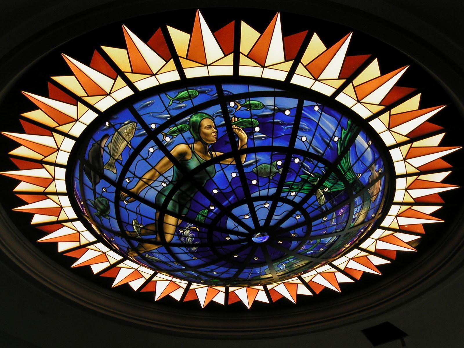 Stained glass dome featuring a central figure swimming amidst blue water and abstract stars, framed by orange rays.