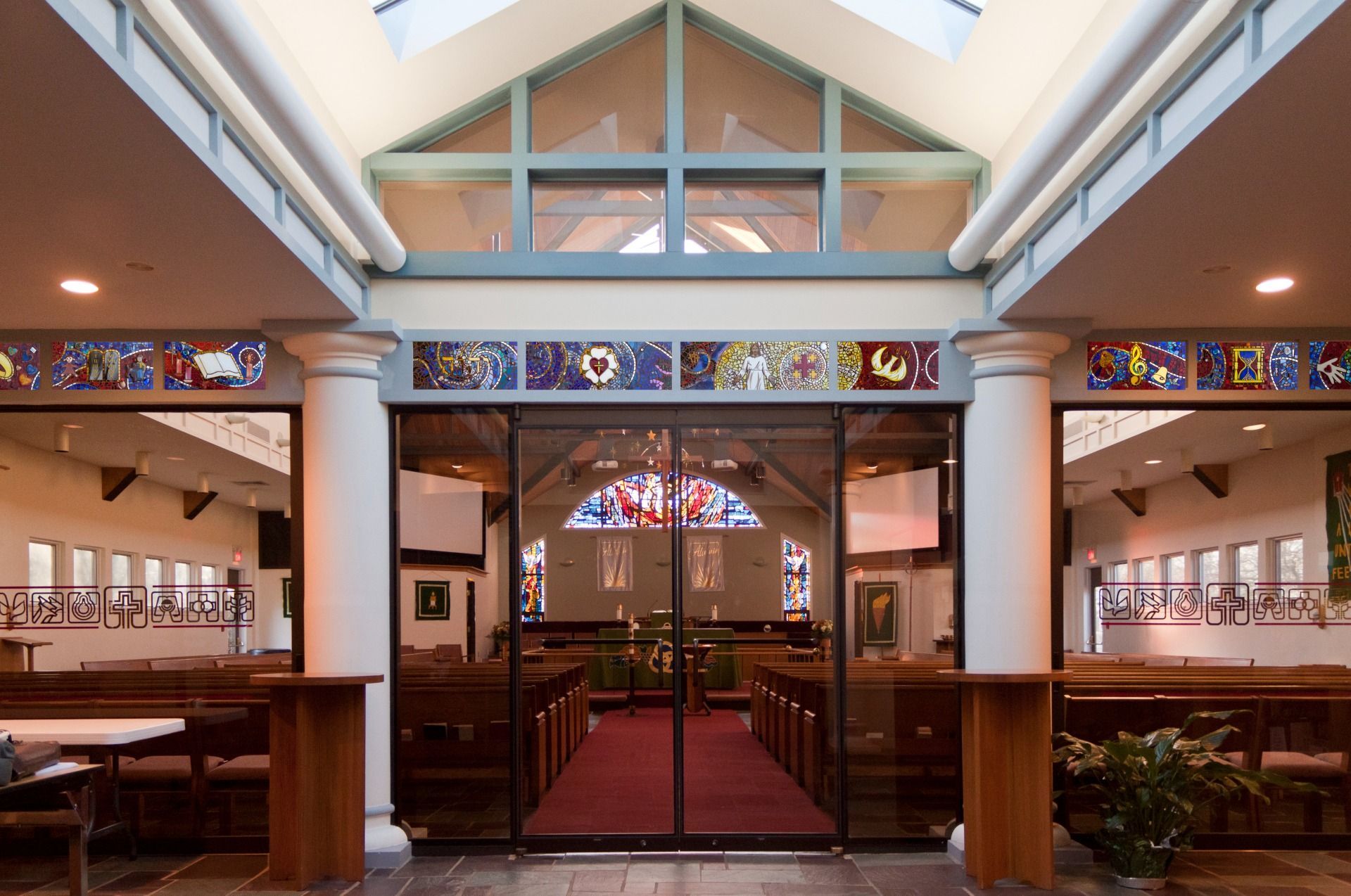Interior view of a modern church sanctuary with wooden pews, a red carpet, glass walls, and stained-glass accents.