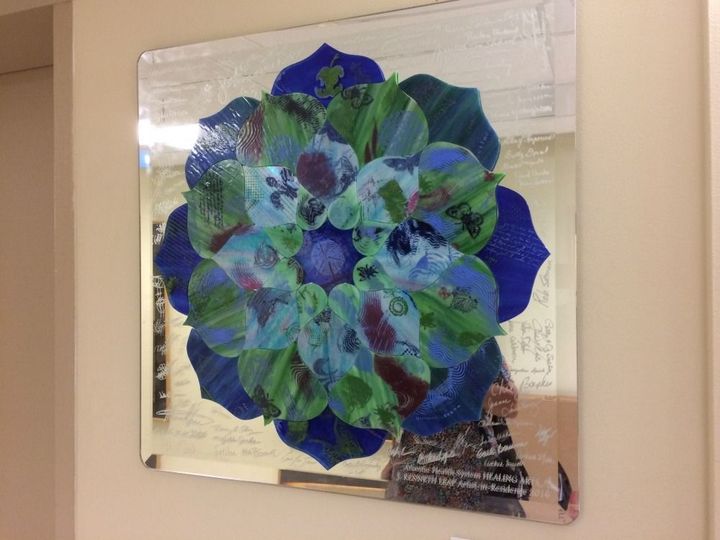 A vibrant, mandala-style collage of blue and green leaves arranged in a circular pattern on a square reflective mirror.