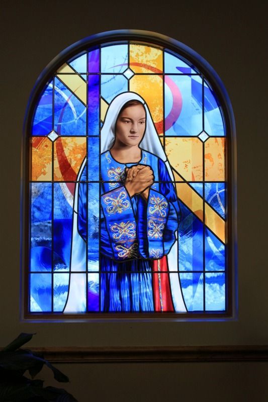 Stained-glass Madonna in blue robe with hands clasped, set in an arched church window.