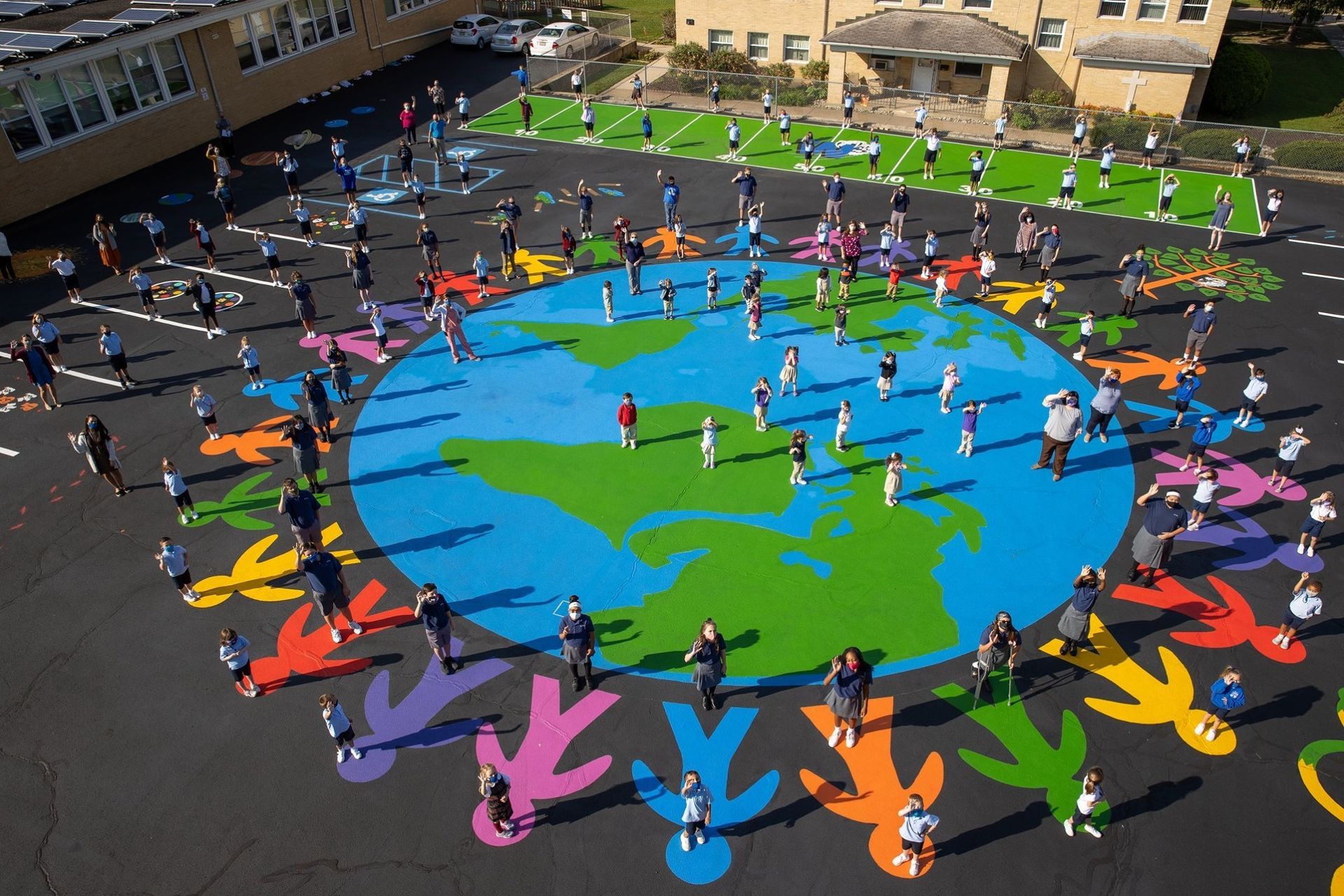 Children stand on a painted playground mural of the Earth surrounded by colorful, painted human figures in a circle.