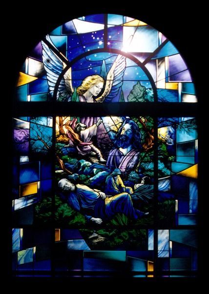 Stained glass window depicting an angel comforting a figure in a garden at night, surrounded by sleeping men.