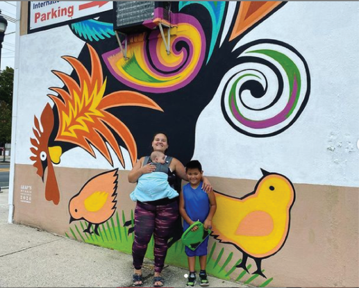 A person carrying a baby in a carrier and a child stand in front of a colorful wall mural featuring a large rooster.