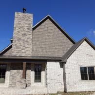 Brick house exterior with stone chimney, dark gray gabled roof, and white siding under a blue sky