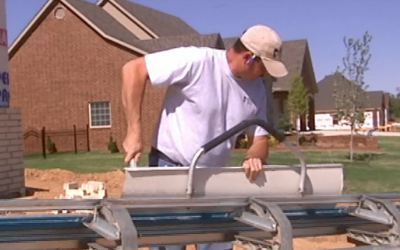 Worker in white shirt and cap adjusts a machine outdoors beside a brick house.