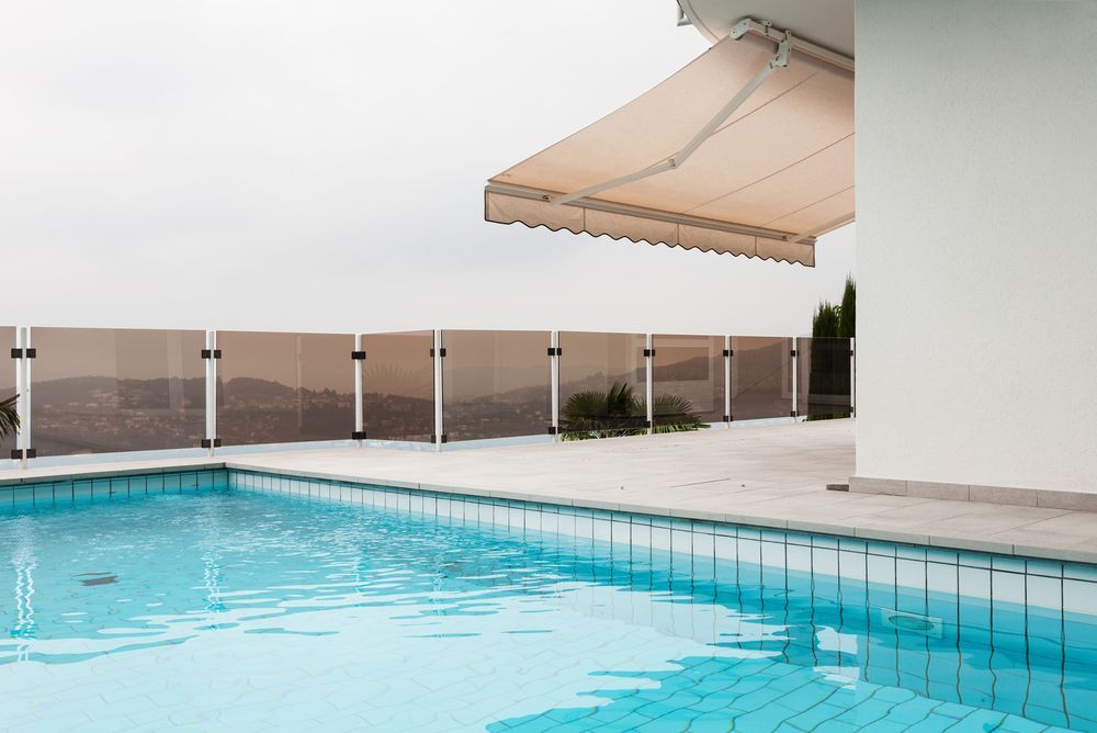 Modern poolside area with clear blue water, glass fencing, and a beige retractable awning extending 