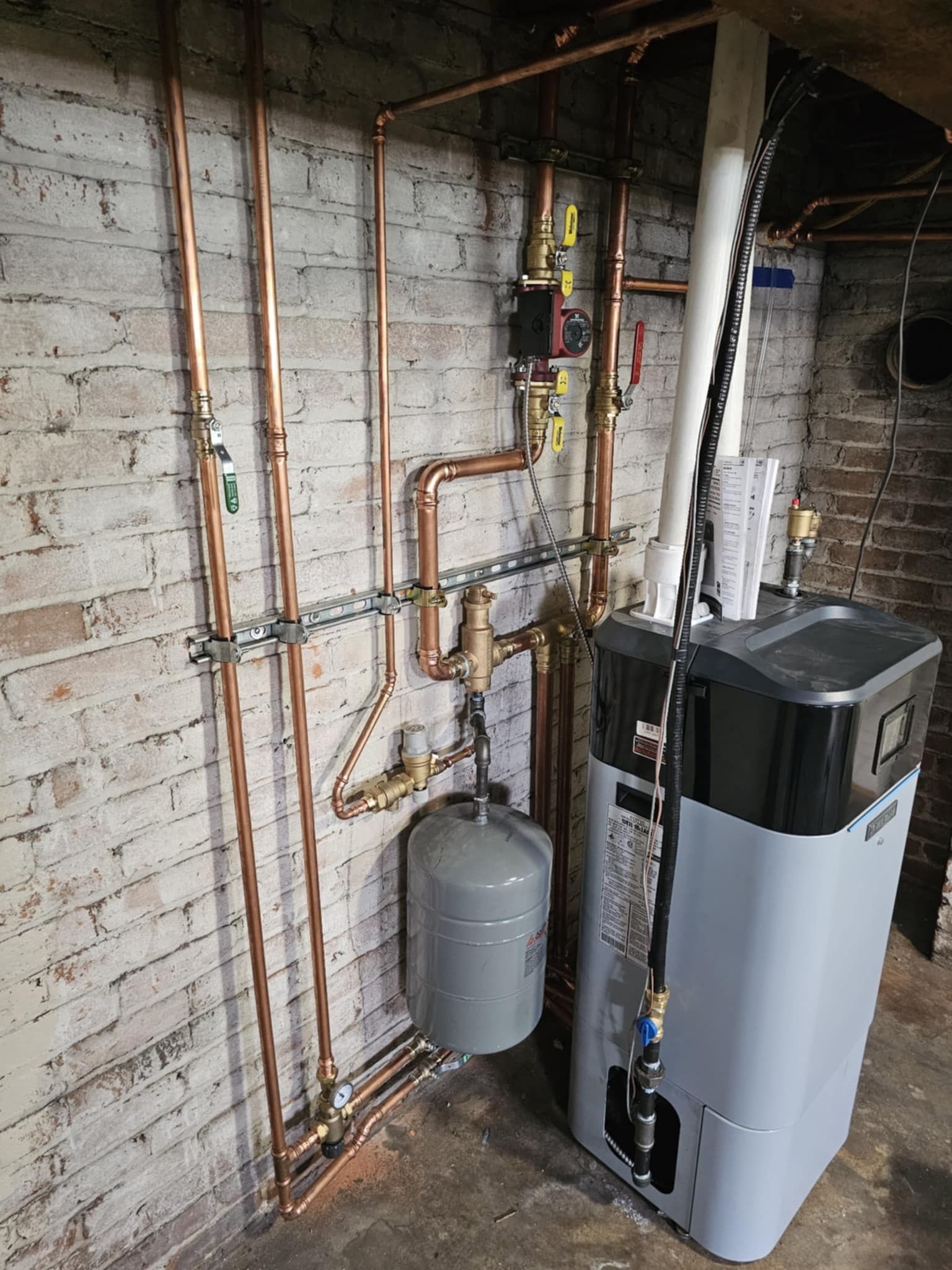 A water heater is sitting in a basement next to a brick wall.