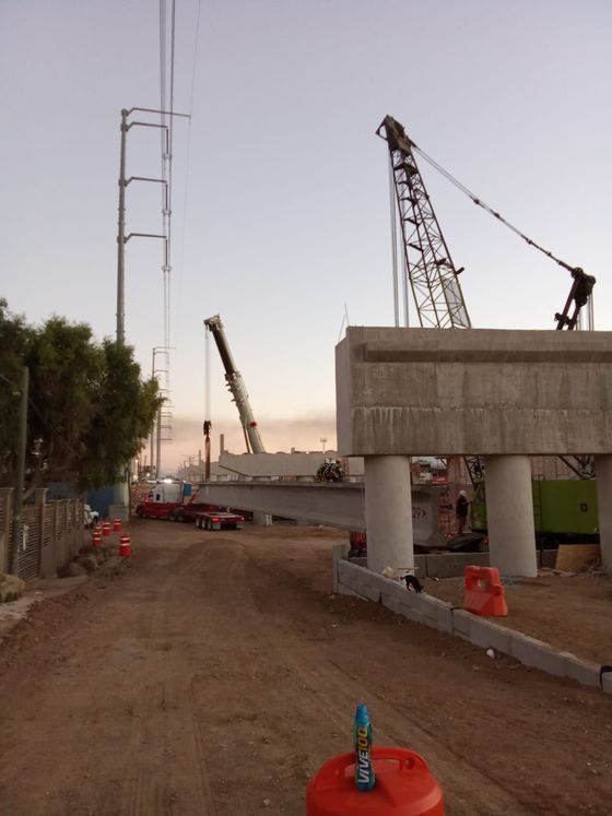 Sitio de construcción con grúas levantando secciones de puentes de hormigón; líneas eléctricas al fondo.