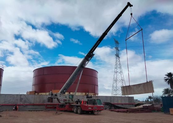 Una grúa levanta un encofrado de hormigón rectangular cerca de un gran tanque de almacenamiento rojo bajo un cielo nublado.