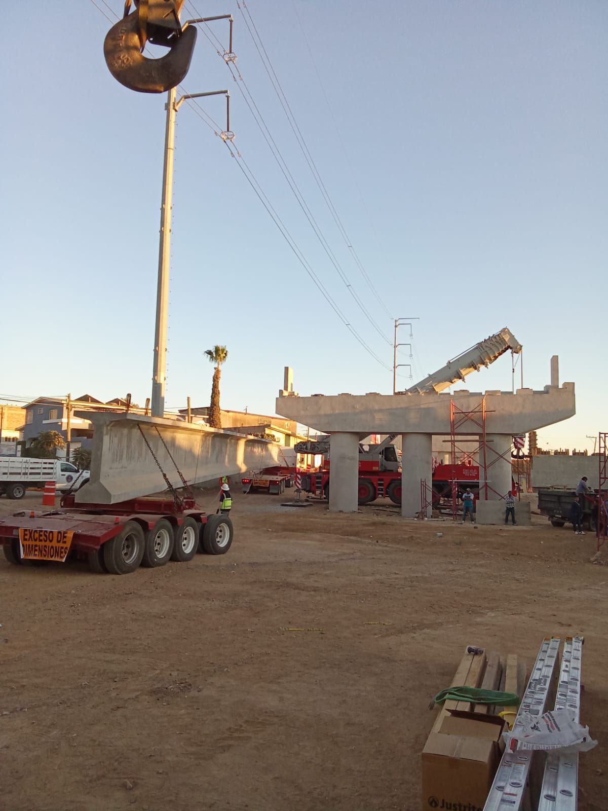 Sitio de construcción: Grúa levantando sección de puente de concreto cerca de líneas eléctricas y soportes.