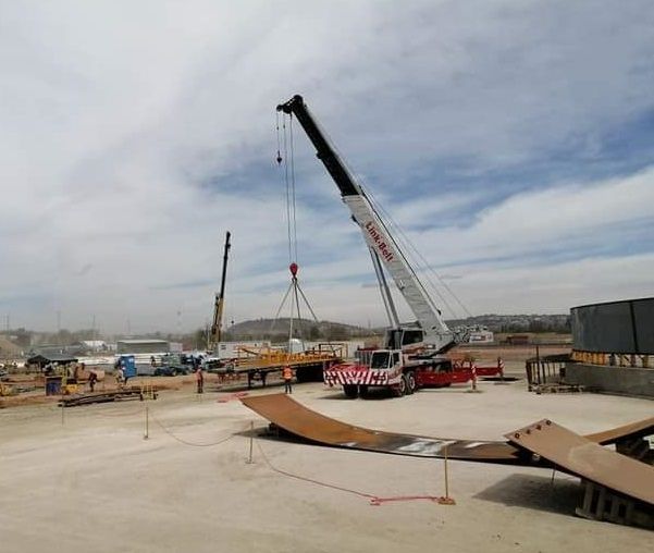 Una gran grúa levanta una viga de metal en un sitio de construcción en un día nublado.