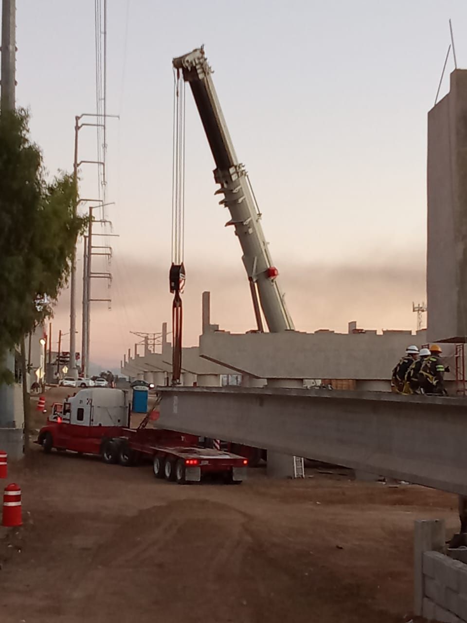 Una grúa levanta una viga de hormigón durante la construcción de una carretera. Un camión transporta la viga, con una puesta de sol de fondo.