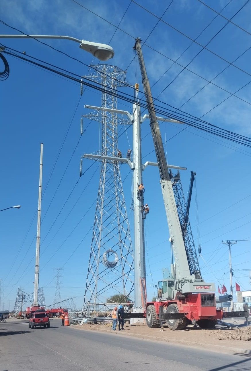Una grúa levanta una torre de tendido eléctrico en un día despejado y soleado. Hay trabajadores en la torre.