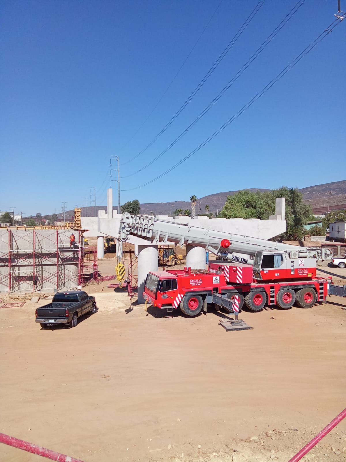 Sitio de construcción con una gran grúa, tubería, camiones rojos y un automóvil pequeño bajo un cielo azul.