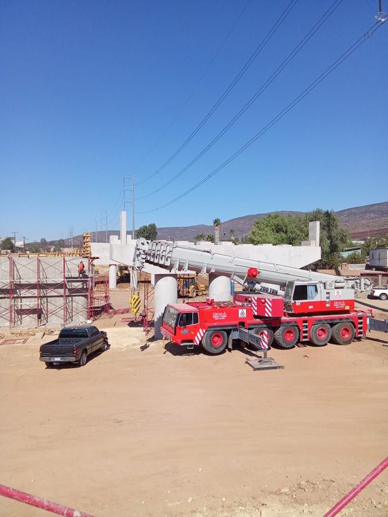 Sitio de construcción con una gran grúa, tubería, camiones rojos y un automóvil pequeño bajo un cielo azul.
