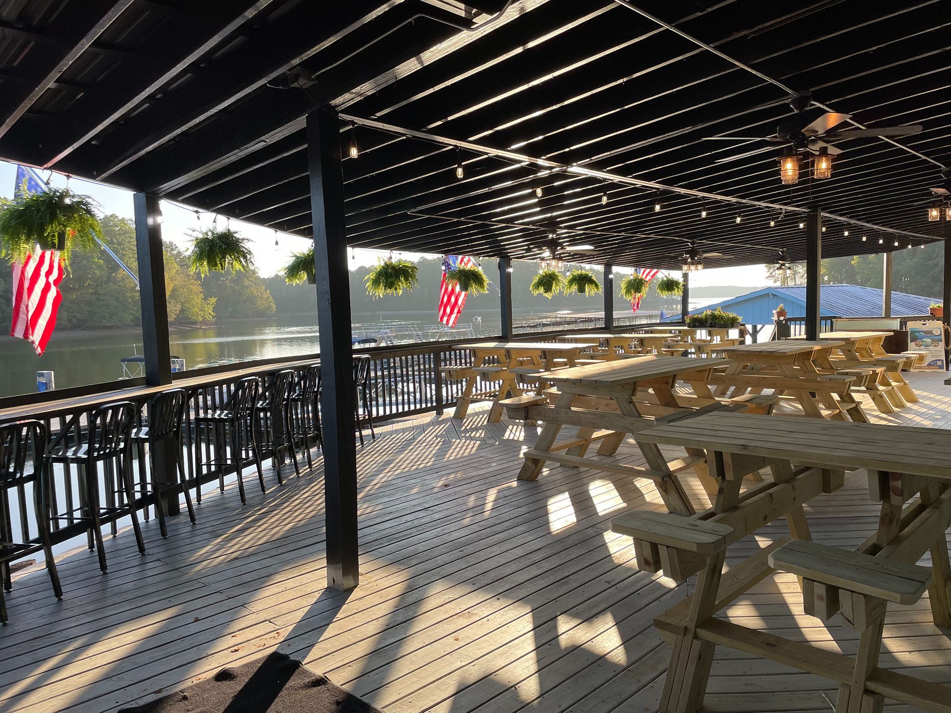 Outdoor dining area with picnic tables on a wooden deck overlooking water; string lights and hanging plants.