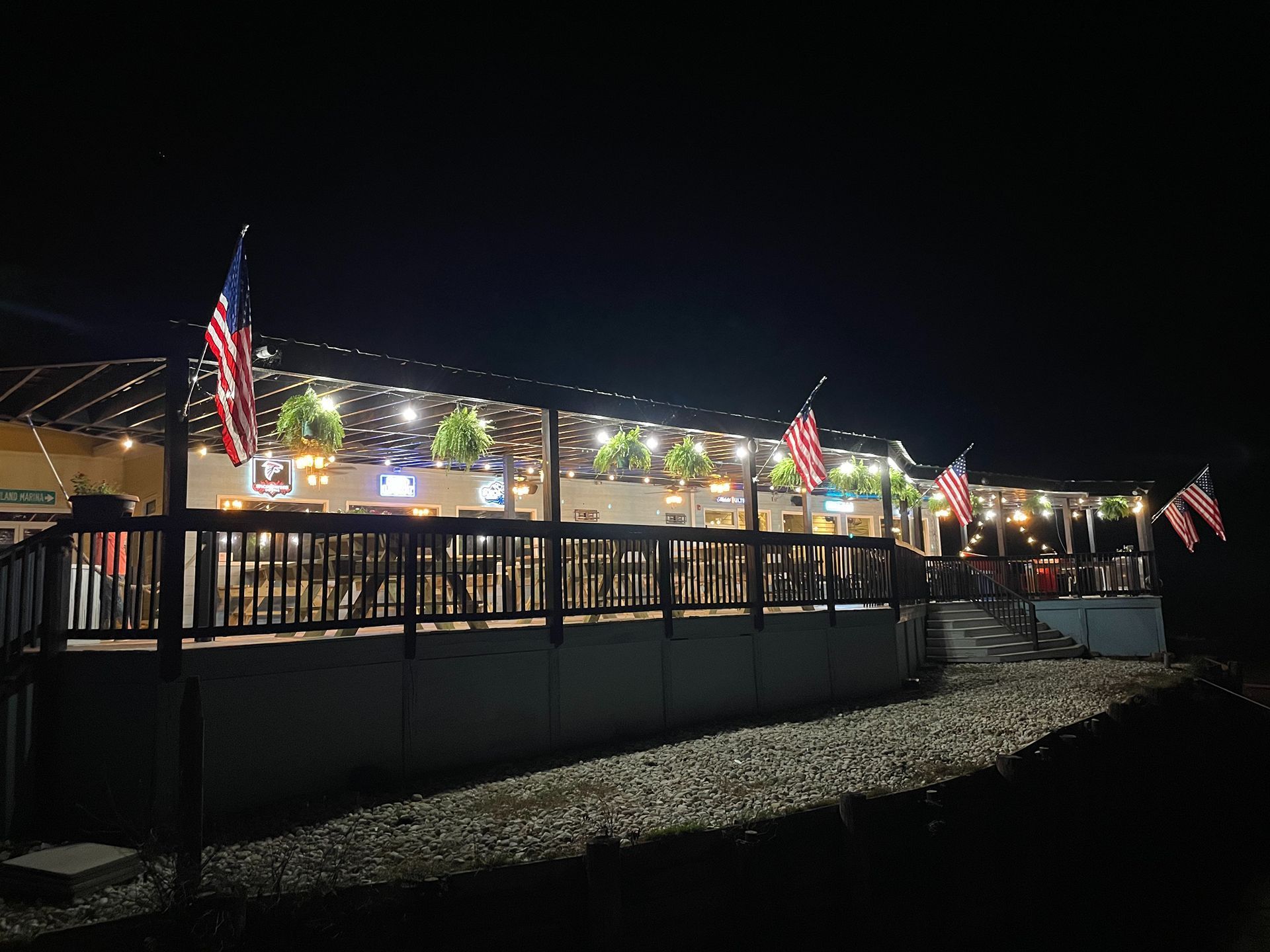 Nighttime view of a restaurant patio with American flags and string lights; people are visible at tables.